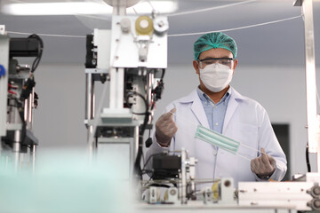 Worker man in personal protective equipment or PPE inspecting quality of mask and medical face mask production line in factory, manufacturing industry and factory concept.