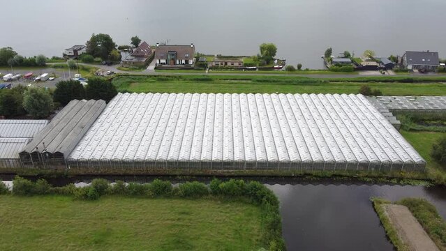 Drone aerial shot of a large european greenhouse with a lake and a town in the background. A home and a few cars surround the greenhouse.