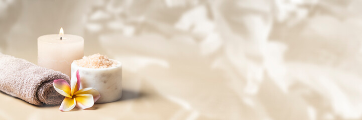 Obraz na płótnie Canvas Beauty Spa salon banner. Burning candle, towel with frangipani flower and salt scrub on beige background with tropical leafe texture.