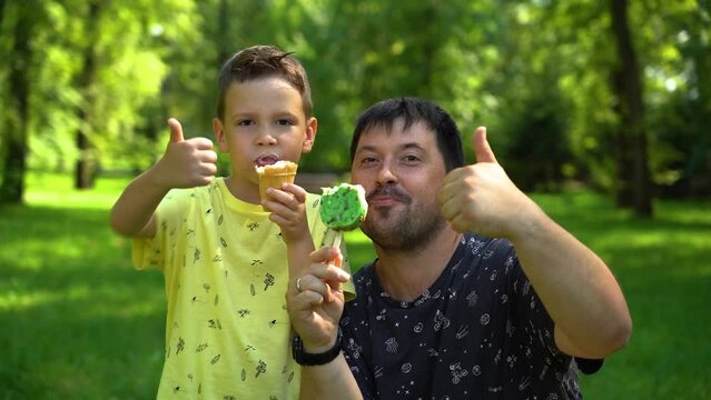 Father and son eat ice cream and gesture cool on a sunny summer day in the park to satisfy their hunger and cool down. Boy and dad eat delicious ice cream in the park among green trees