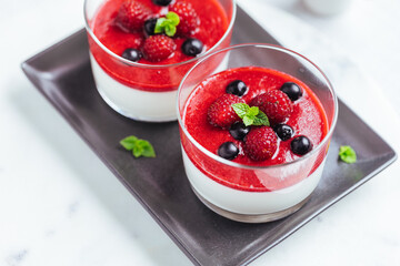 Panna cotta dessert with sauce from berries. Cherries, raspberries and blueberry as decoration. Served in two glasses on dark background. Traditional Italian creamy dessert.
