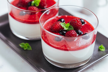 Panna cotta dessert with sauce from berries. Cherries, raspberries and blueberry as decoration. Served in two glasses on dark background. Traditional Italian creamy dessert.