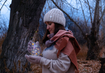 A girl in a knitted hat holds a jar with a garland in her hands, admires the flickering of lights.
