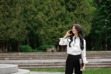 A pretty woman in a white shirt stands in the park with a folder in hand talking on a smartphone.