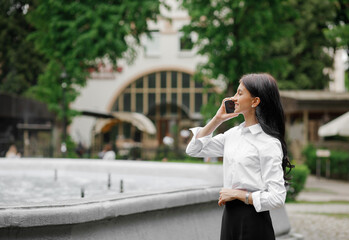 Fototapeta premium Beautiful, caucasian girl talking on the phone on the background of a fountain in the park. Freelance concept.