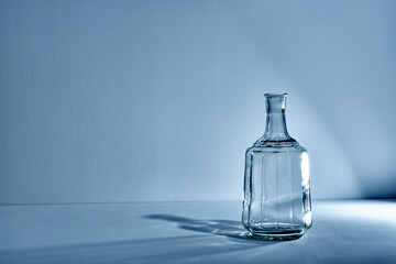 Glass empty open bottle with copy space. Graphic still life with light and shadow in blue light. The concept of glass containers, recyclable materials or alcoholism. Large creative vase. Open flask