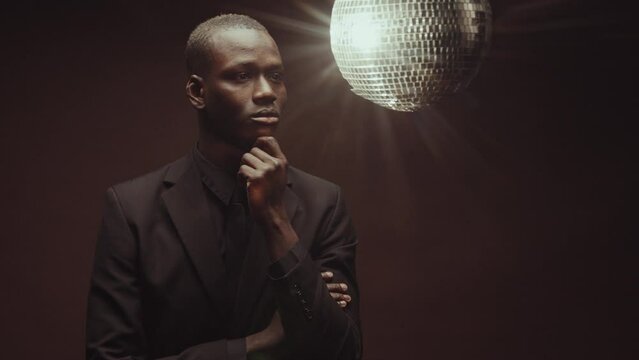 Conceptual medium length portrait of handsome young adult African American man wearing elegant black suit thinking of something, disco ball rotating on background