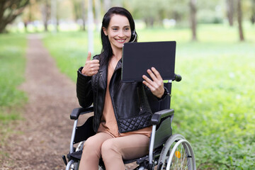 Smiling woman sitting in wheelchair in park and showing thumb up on tablet of camera