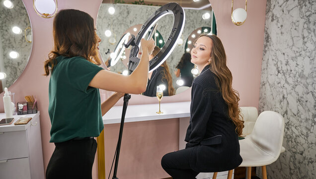 Makeup artist photographing client in beauty salon. Young woman with long hair sitting on chair and looking at beauty specialist while female worker taking picture with smartphone.