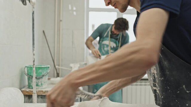 Selective focus of young adult Caucasian man wearing apron working with plaster cast removing extra layers of material using hand tool, prosthetic production routine