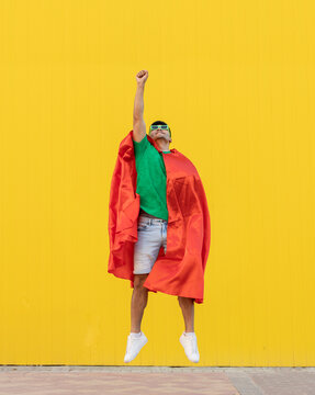 Young man wearing red cape jumping in front of yellow wall