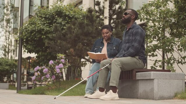 Slowmo of young curvy African American woman reading book to blind man with walking stick, sitting together on bench in park