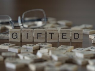 Gifted word or concept represented by wooden letter tiles on a wooden table with glasses and a book - obrazy, fototapety, plakaty
