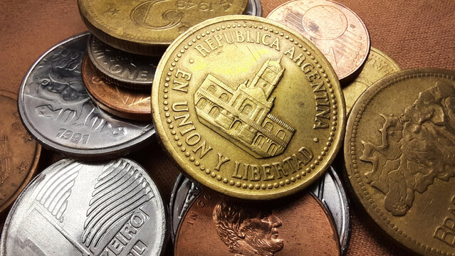 Golden Peso coin, with the image of the Cabildo of Buenos Aires building, together with a collection of various old coins on a brown background.
