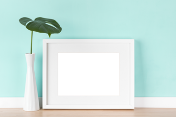 Elegant horizontal transparent white picture frame with matte and monstera leaf in vase in front of pastel green wall.