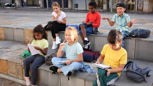 Positive preteen classmates preparing schoolwork, sitting on steps outside school in sunny spring day
