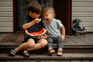 Two brothers sit on the porch of a village house and eat watermelon with a spoon, an older boy...