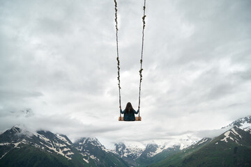 A young woman rides on a swing against the backdrop of snowy rocky mountains. Extreme entertainment in the mountains of the Caucasus
