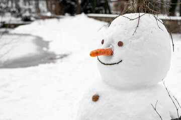 Old funny fat snowman with nose carrot nose, buttons made of walnuts, cork eyes stands alone in open snow-covered field near frozen lake in park on quiet winter december, january or february day