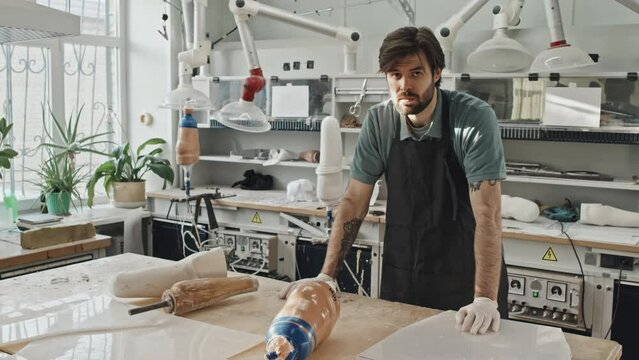Portrait of young adult Caucasian man with beard on face wearing apron and latex gloves standing at workbench in prosthetic production workshop looking at camera