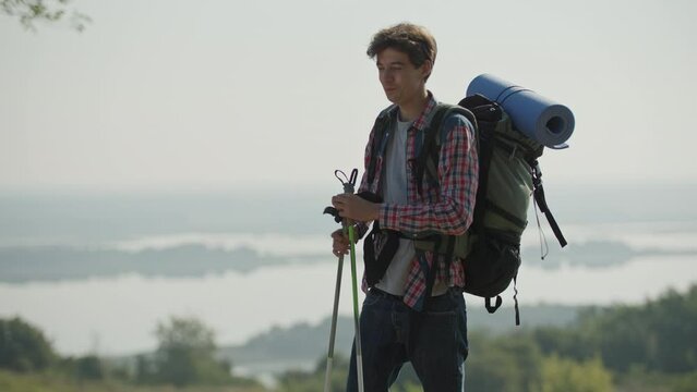 20s Man Hiking On The Riverside With Tourist Backpack And Trekking Poles Early In The Morning. Person Looking For Route Using Smartphone.