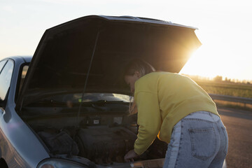 a girl with blond hair in casual clothes stands with an open hood at sunset and looks at what has...