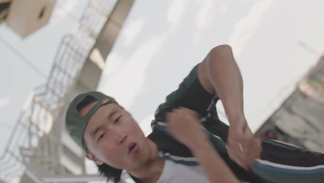Slowmo of Asian guy wearing backwards baseball cap hip hop dancing outdoors at camera