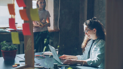 Two attractive girls are working together in modern loft office indoors Curly businesswoman is typing on laptop sitting at table while her colleague is reading book standing near window.