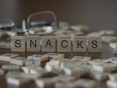 Snacks word or concept represented by wooden letter tiles on a wooden table with glasses and a book