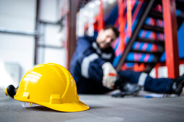 Safety at work. Close up view on hardhat and injured factory worker in background. - obrazy, fototapety, plakaty
