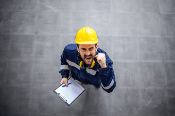 Top view of happy factory worker in protective work wear and hardhat celebrating job results, promotion and success.