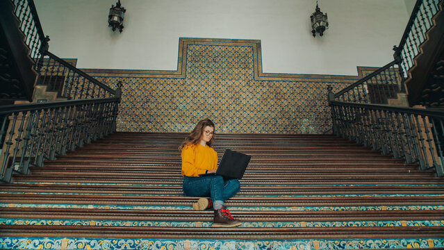 Cinematic symmetric shot of young woman in yellow sweater sit on stairs of old university campus building, work on laptop on new exciting school project or assignment. Freshmen or junior student
