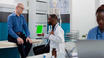 African american medic pointing at tablet with greenscreen display, looking at chromakey template with old patient. Physician and man using isolated mockup background and blank copyspace.