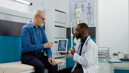 Obraz premium Medic physician explaining x ray scan results to patient in medical cabinet, analyzing bones radiography diagnosis on digital tablet to give treatment. Healthcare checkup appointment with doctor.