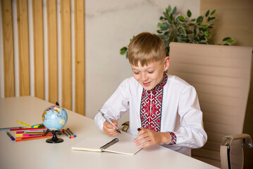 schoolboy in ukrainian embroidery writing in notebook and sitting at the desk at school with globe and different colored penciles - obrazy, fototapety, plakaty