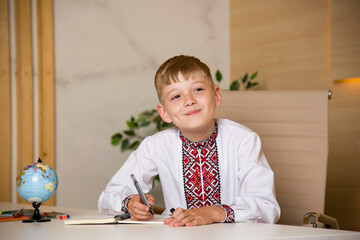 Fototapeta na wymiar schoolboy in ukrainian embroidery writing in notebook and sitting at the desk at school with globe and different colored penciles