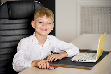 smiling boy with glasses sitting in the director's chair in the office of the school with laptop