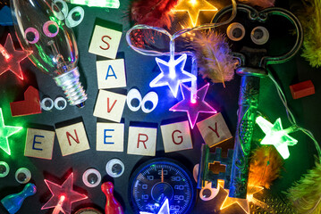 The words save and energy laid with wooden stones surrounded by eyes, an incandescent lamp, a string of lights, an old key, a thermometer and colorful feathers to draw attention to saving energy 