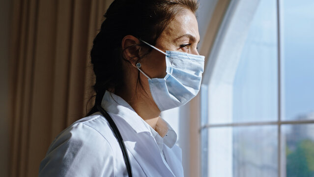 In front of the camera mature woman very concentrated and with thinking face take off her mask off while have a break and looking through the panoramic window in the hospital