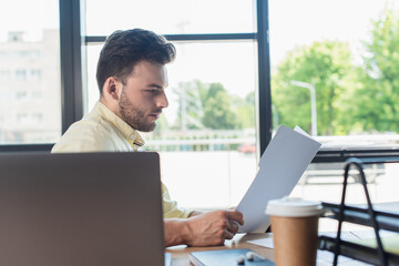 Side view of businessman holding paper folder near blurred laptop and coffee to go in office.