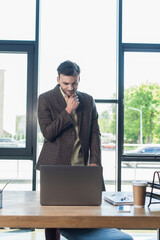 Smiling businessman in jacket looking at laptop near notebook and paper cup in office.