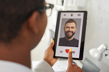 Young woman finds her love online. Happy African American girl gives like to picture of smiling handsome Caucasian man on modern mobile love app or dating website. Close up of hands and tablet screen