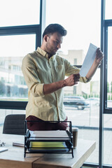 Businessman looking at document near coffee to go and papers in office.