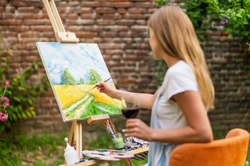Woman enjoys painting on canvas and drinking wine outdoor.