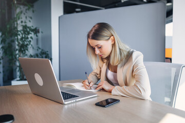 Obraz premium Serious women in classic clothes, writing notes about the training for students, works at the table with a laptop.