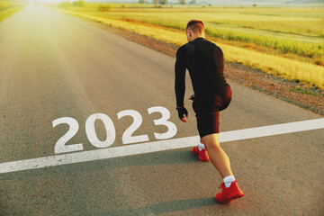 New year 2023 or start straight concept Word 2023 written on the asphalt road and athlete man runner stretching leg preparing for new year at sunset. Concept of challenge or career path and change.