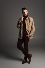 Full length of man in autumn outfit holding paper cup and posing on grey background.