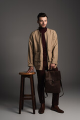Full length of bearded man in autumn outfit holding backpack near chair on grey background.