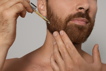 Man applying oil onto beard on grey background, closeup