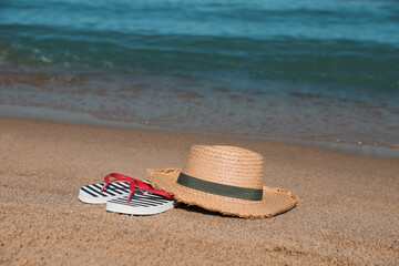 Stylish straw hat and striped flip flops on sandy beach near sea